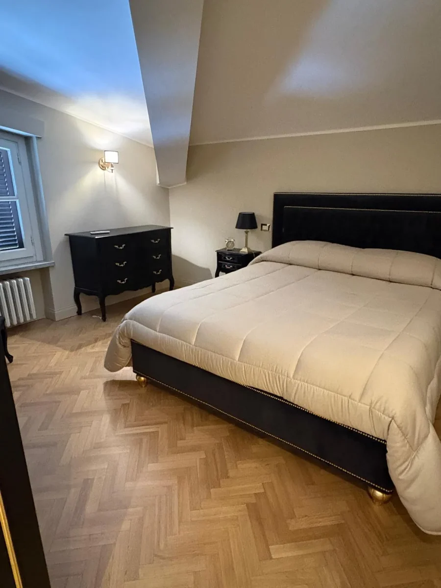 Veduta d'insieme della camera con letto, comò laccato nero, applique e parquet a spina di pesce
