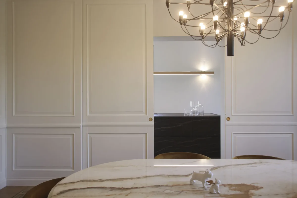 Nicchia bar in legno scuro incassata nella boiserie bianca, con lampadario in primo piano