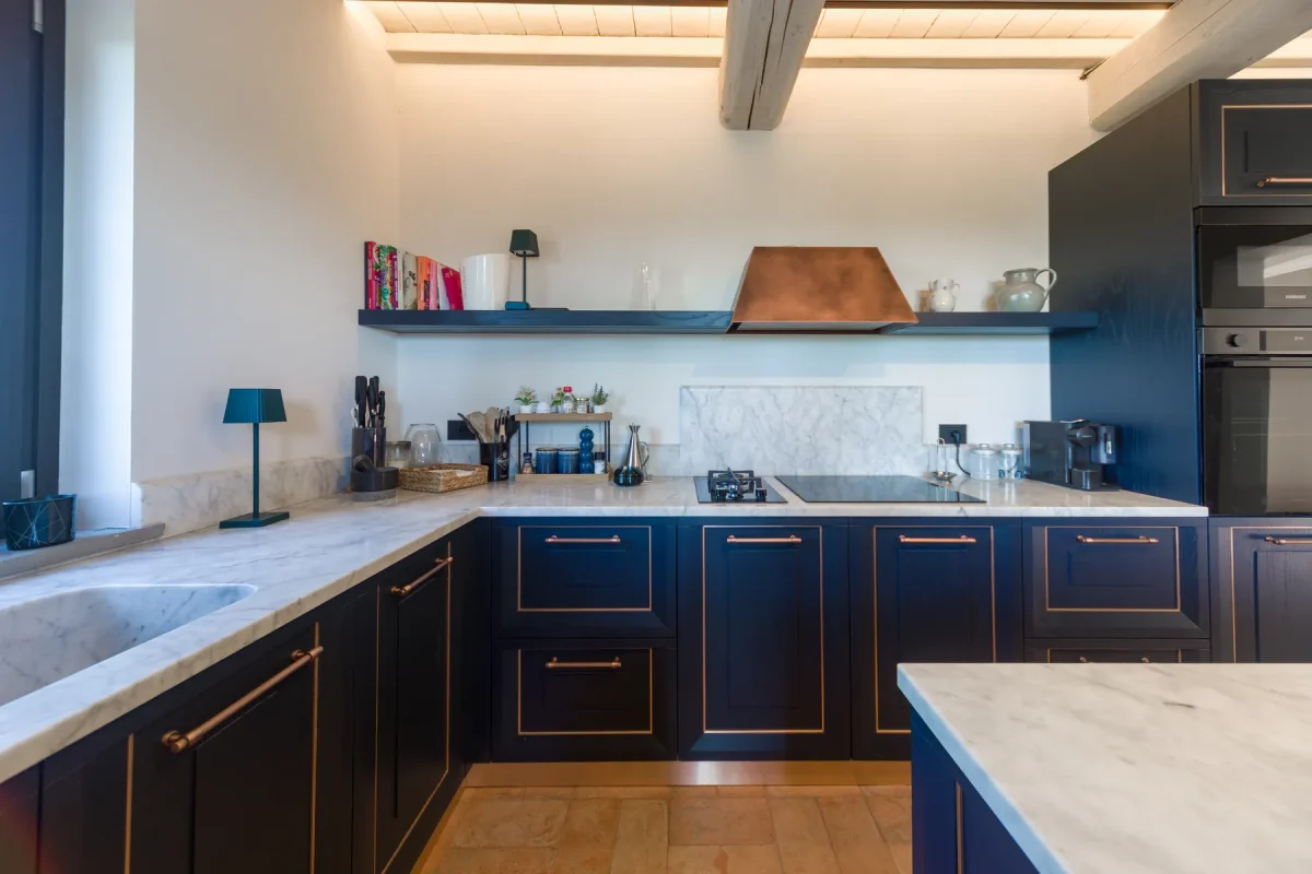 Veduta della cucina a U: basi in rovere laccato blu, piano in marmo di Carrara, cappa in rame e pavimento in cotto sotto le travi imbiancate