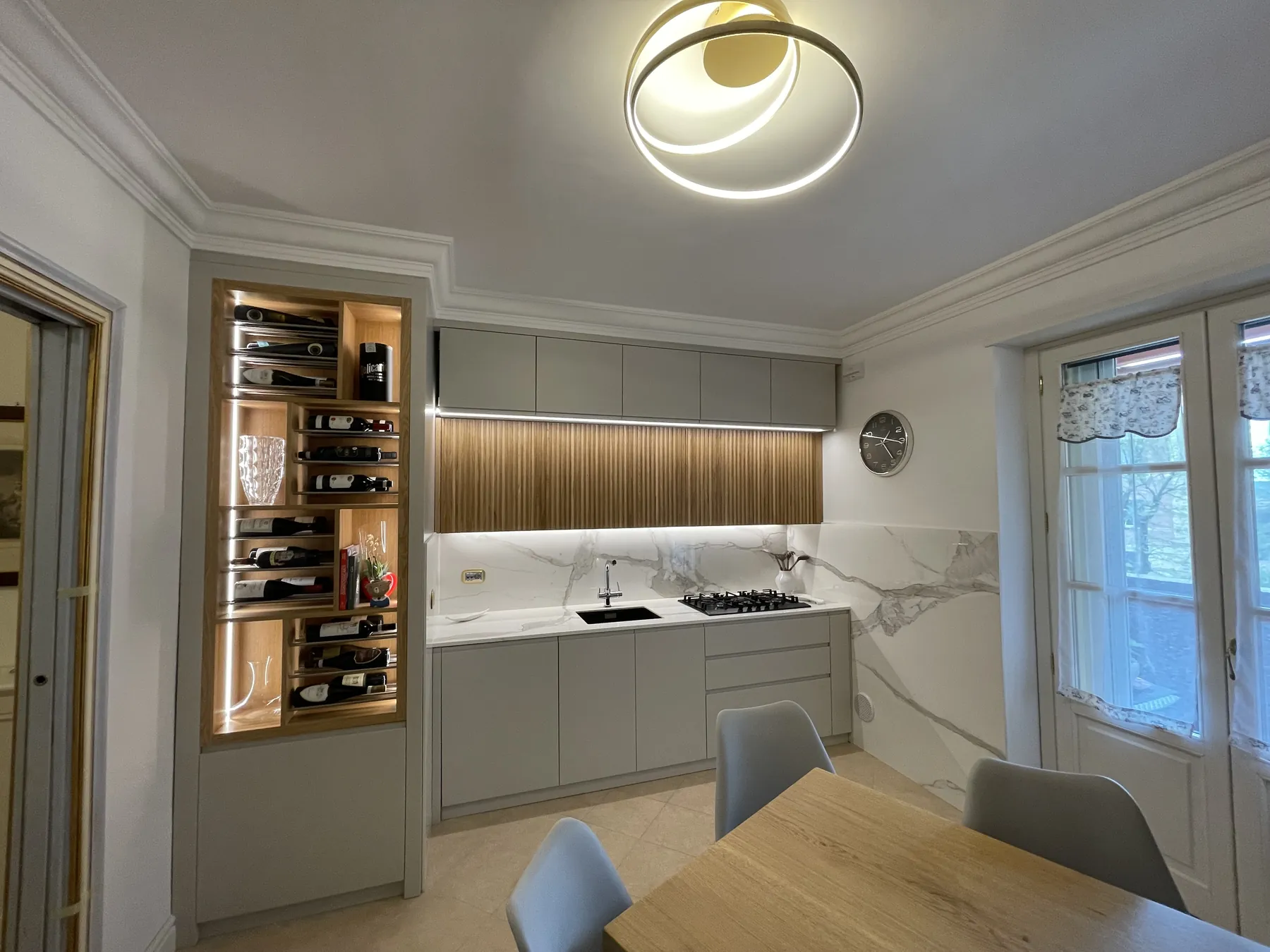 Veduta d'insieme della cucina: nicchia porta-vini illuminata, boiserie in rovere doghato, marmo calacatta e lampada LED circolare