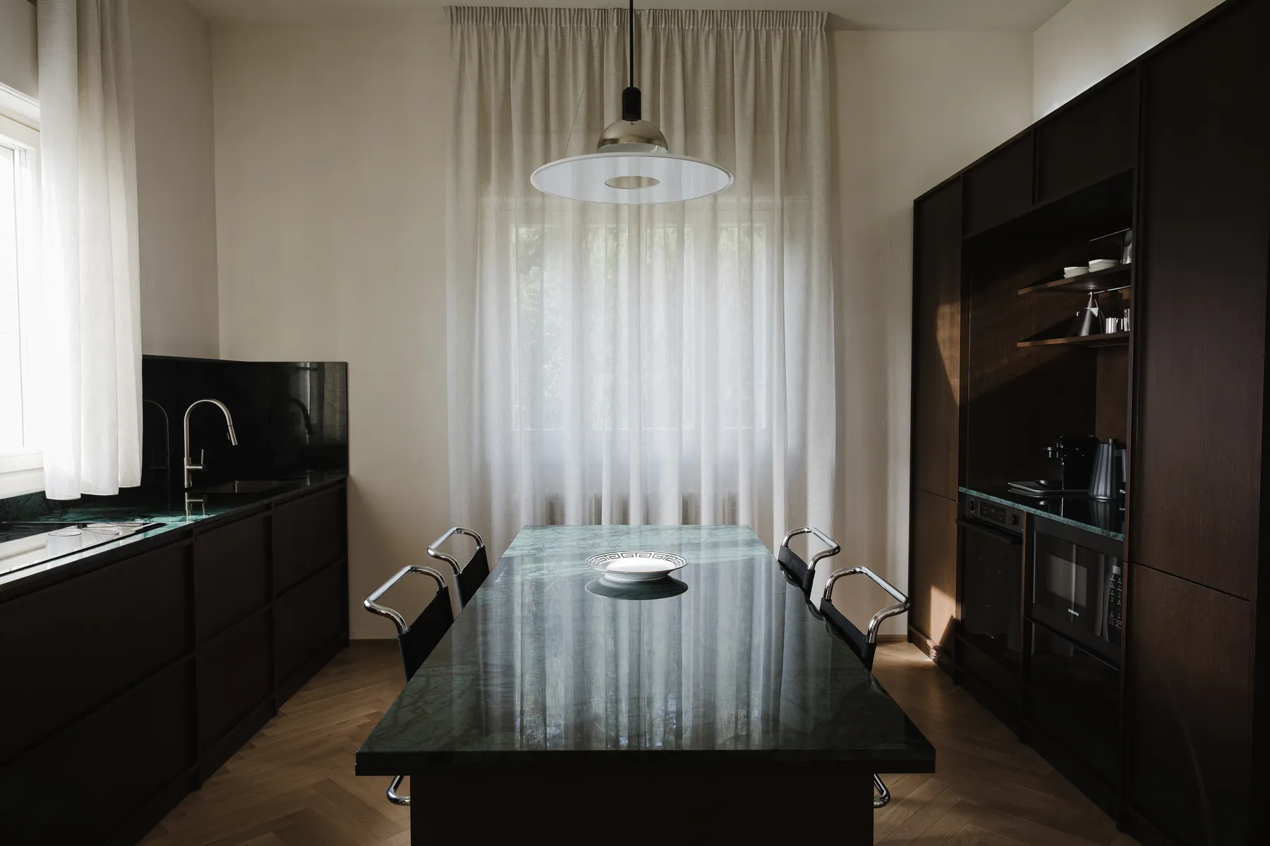 Isola in marmo verde alpi al centro della cucina, con credenza in legno wengè e piani lungo la parete, sotto la luce delle tende a velo.