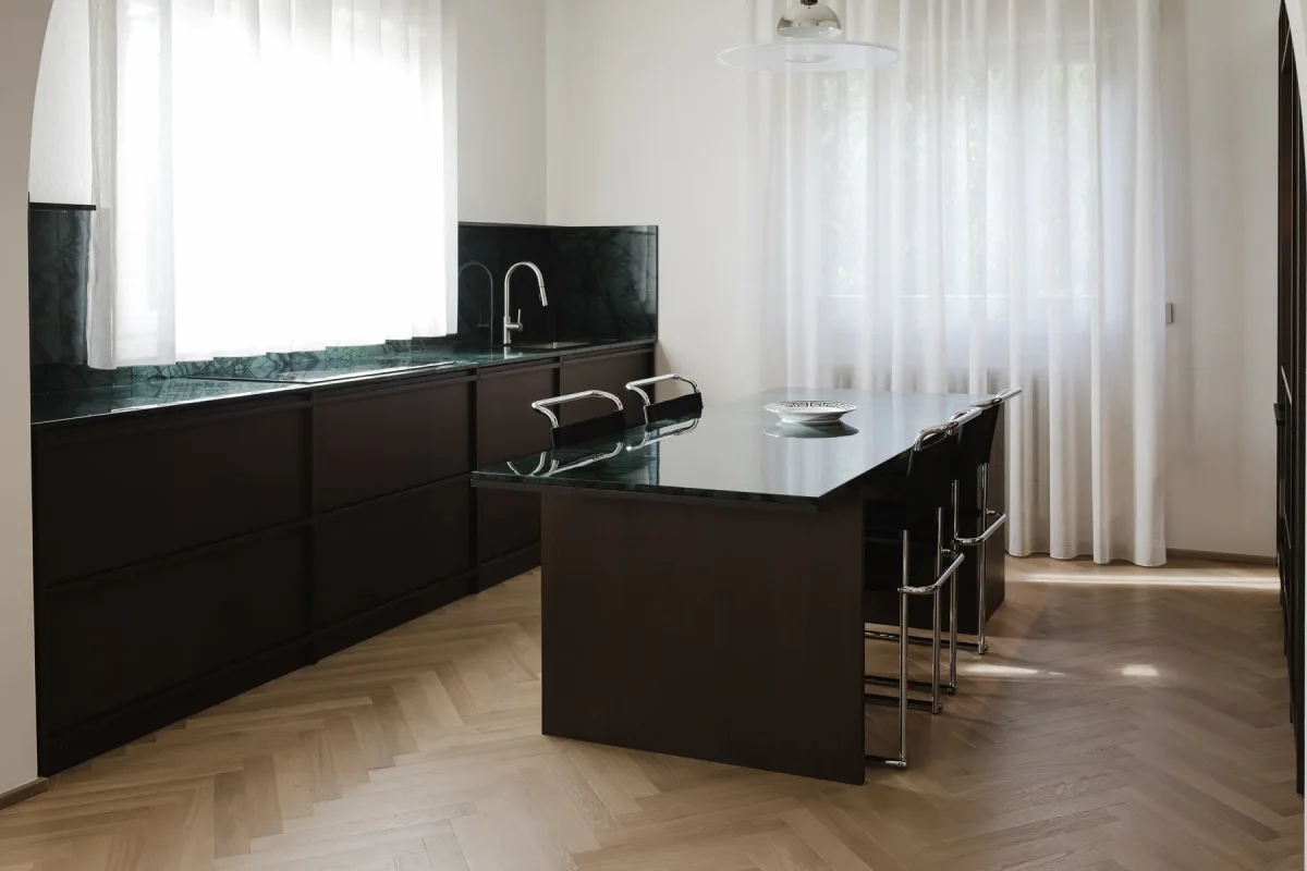 Veduta d'insieme della cucina: piani in marmo verde alpi lungo la parete e isola centrale in legno wengè, su parquet a spina di pesce.