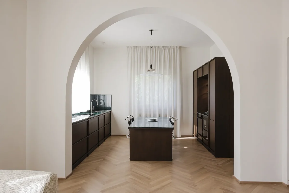 La cucina vista attraverso un'arcata a tutto sesto: l'isola in wengè e marmo al centro, i piani sui lati, il parquet a spina di pesce.