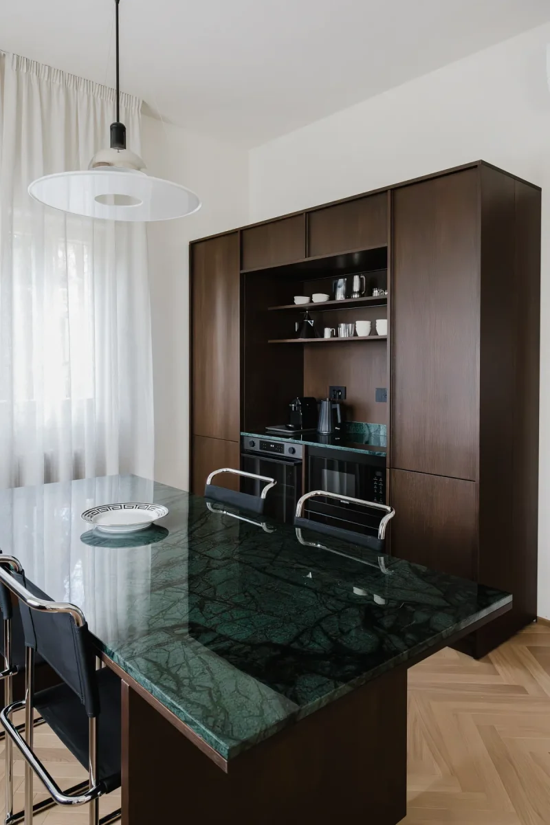 Isola in marmo verde alpi con sedie a sbalzo e piatto a greca; alle spalle la credenza in legno wengè.